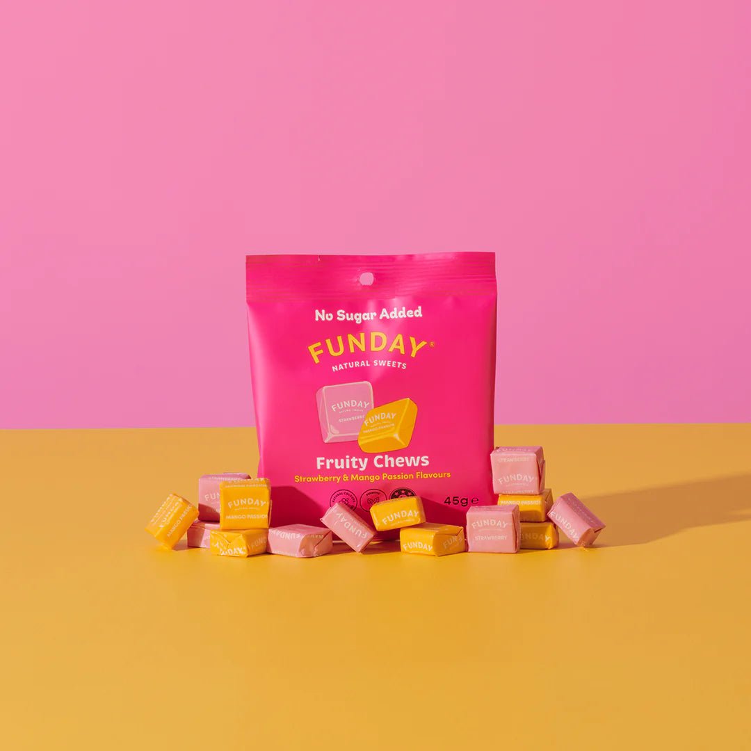 A bright pink Funday Fruity Chews - 45g packet, made with plant-based prebiotic fibre and no added sugar, sits on a yellow surface surrounded by individually wrapped yellow and pink candies, against a pink background.