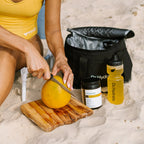 Person slicing a mango on wooden board next to Dr Hydrate Mango Passionfruit Jar - 30 Serves and branded water bottle on sand