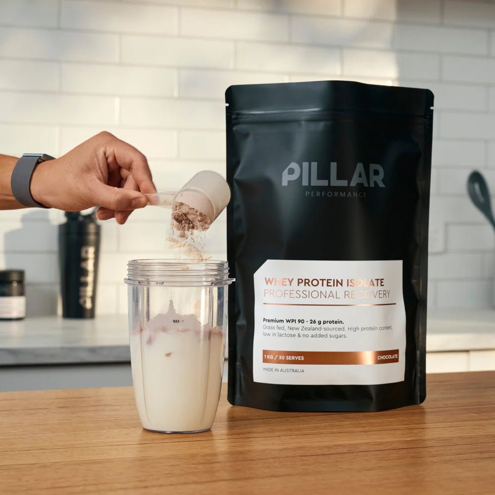 WPI - Chocolate - 1kg by PILLAR PERFORMANCE bag beside a hand scooping protein powder into a blender cup